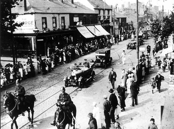 B+W photo of George Inn Public House and High Street 1900s showing a procession of cars led by two horse mounted police officers people line the route