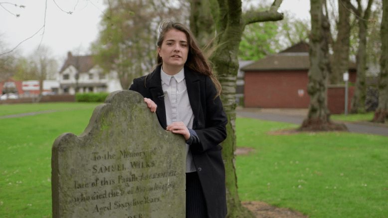 A video still of female student stood by gravestone of Samuel Wilkes